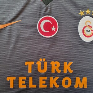 Puede incluir: Camiseta deportiva gris con el logo naranja de Nike, parche de la bandera turca y el emblema del Galatasaray. La camiseta presenta el texto "T&Uuml;RK TELEKOM" en letras naranjas. Tres estrellas doradas est&aacute;n encima del emblema del Galatasaray.