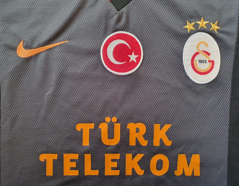 Puede incluir: Camiseta deportiva gris con el logo naranja de Nike, parche de la bandera turca y el emblema del Galatasaray. La camiseta presenta el texto "T&Uuml;RK TELEKOM" en letras naranjas. Tres estrellas doradas est&aacute;n encima del emblema del Galatasaray.
