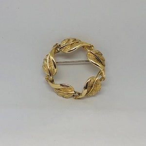 Puede incluir: Un broche dorado en forma de círculo, hecho de hojas enlazadas.