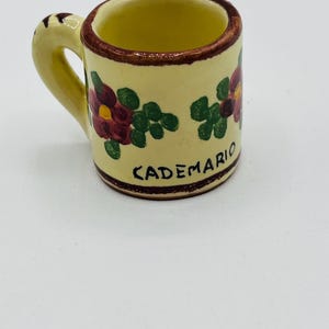 Może przedstawiać: Mały, żółty kubek ceramiczny z brązowym uchwytem i kwiatowym wzorem. Na kubku widnieje napis "CADEMARIO".