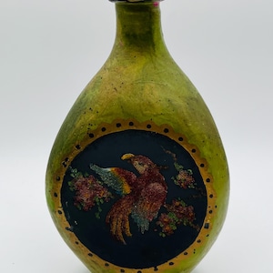 May include: A hand-painted, pear-shaped bottle with a green and gold finish. A black circular design features a colourful bird and floral accents. The bottle has a pink and black rim.
