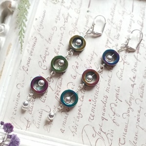 May include: A pair of dangle earrings with iridescent, circular beads in shades of green, blue, and purple. The earrings are connected by silver wire and feature a small silver bead at the bottom. The earrings are displayed on a white framed background.