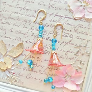May include: A pair of handmade earrings with gold-colored hooks. Each earring features a translucent, bell-shaped, coral-colored bead with gold accents, adorned with blue crystal beads. The earrings are set against a backdrop of vintage paper and floral accents.