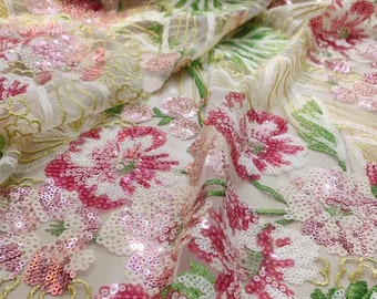 May include: Close-up of a sheer fabric with a floral design. The fabric features large pink and white flowers with sequin details, green leaves, and gold embroidery. The background is a soft, off-white color.