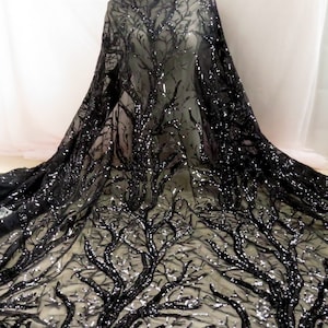 May include: Black sequined fabric featuring a tree branch design. The fabric is draped, highlighting the detailed pattern of branches and sparkling sequins. The background is a soft, neutral colour.