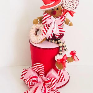 May include: A festive Christmas decoration featuring a gingerbread man wearing a red top hat, holding a lollipop and a donut. The decoration is embellished with red and white striped bows and candy canes, creating a cheerful holiday display.
