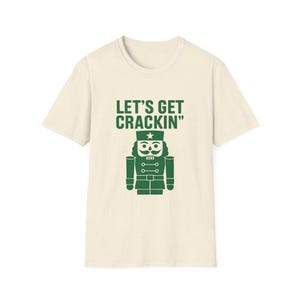 May include: A cream-coloured t-shirt featuring a green nutcracker graphic and the text "LET'S GET CRACKIN'". The nutcracker has a star on its hat and a detailed uniform. The shirt is made of a soft material.