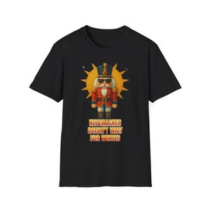 May include: A black short-sleeve t-shirt featuring a graphic of a nutcracker wearing sunglasses in front of a yellow sunburst. Below the nutcracker, the text reads: NUTCRACKER DOESN'T WAIT FOR WINTER in a bold, orange and yellow gradient font.