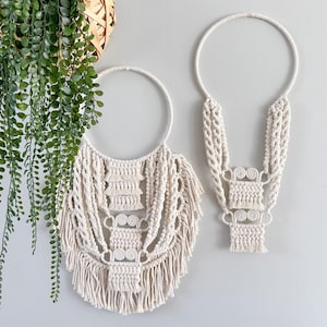 May include: Two off-white macrame wall hangings. The larger one has a circular top with intricate knotting and fringe. The smaller one has a similar design, with a circular top and a vertical pattern. Both are displayed against a light gray wall.