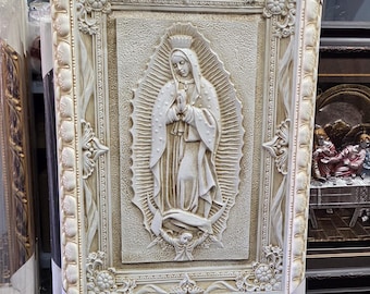 May include: A rectangular wall plaque featuring a relief sculpture of the Virgin of Guadalupe in a cream and gold finish. The central figure is surrounded by a sunburst halo and stands on a crescent moon, all encased in an ornate, decorative frame.