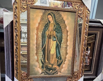 May include: A rectangular wall mirror featuring an ornate gold-colored frame with floral and leaf relief details. The central glass panel displays a printed image of Our Lady of Guadalupe, depicted in a dark green mantle and gold robe, surrounded by rays.