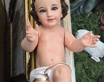 May include: A detailed figurine of a baby with blue eyes and brown hair, wearing a white diaper with gold trim. The figurine is part of a religious display, with other figures and a gold-framed picture in the background.
