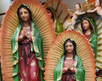 May include: Two Virgin of Guadalupe statues with gold accents. The statues are standing on a cloud with an angel underneath. The statues are made of painted resin and are approximately 12 inches tall. The statues are on a shelf with other religious items.
