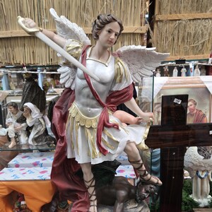 May include: A detailed statue of Saint Michael the Archangel, depicted with wings, a sword, and a defeated demon at his feet. The statue is predominantly white, gold, and red, with intricate detailing. The base of the statue is inscribed with the words "San Miguel".