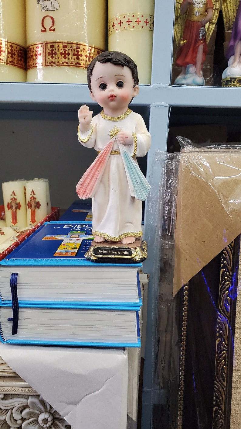 Puede incluir: Estatua religiosa de un ni&ntilde;o peque&ntilde;o con una t&uacute;nica blanca con adornos dorados, levantando una mano. La figura tiene una luz rosa y azul que emana de su pecho. Est&aacute; sobre una pila de libros de tapa azul. La base de la figura dice "Divina Misericordia".