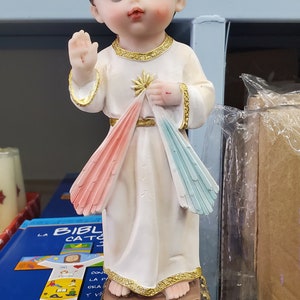 Puede incluir: Estatua de un ni&ntilde;o peque&ntilde;o con una t&uacute;nica blanca con detalles dorados, levantando una mano en se&ntilde;al de bendici&oacute;n. La t&uacute;nica presenta rayos rosas y azules. La base est&aacute; inscrita con "Divina Misericordia".