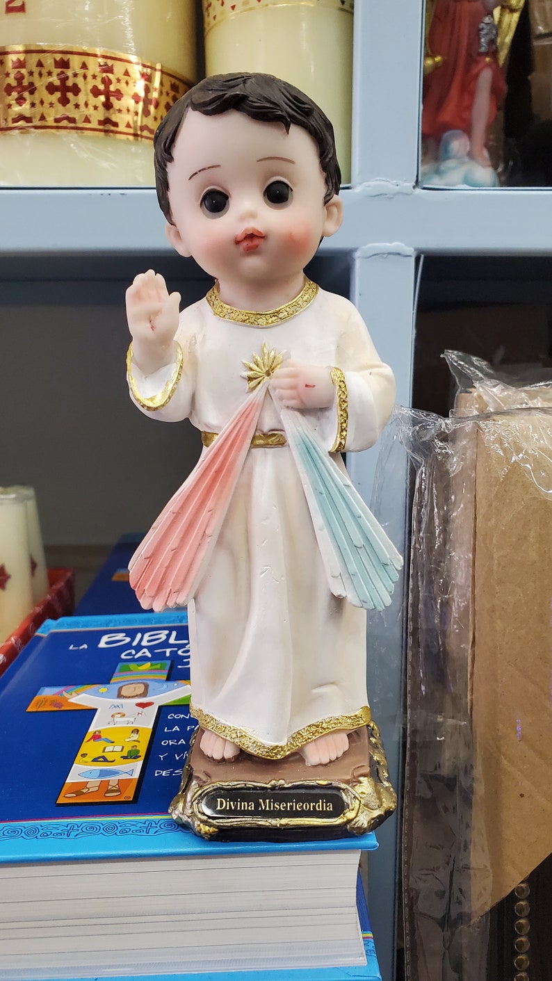 Puede incluir: Estatua de un ni&ntilde;o peque&ntilde;o con una t&uacute;nica blanca con detalles dorados, levantando una mano en se&ntilde;al de bendici&oacute;n. La t&uacute;nica presenta rayos rosas y azules. La base est&aacute; inscrita con "Divina Misericordia".