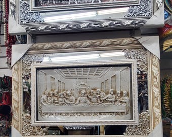 May include: A rectangular wall mirror featuring a central relief sculpture of The Last Supper. The frame is ornate, cream-colored with floral carvings at the corners and a leaf-patterned border. The relief depicts figures seated at a long table.