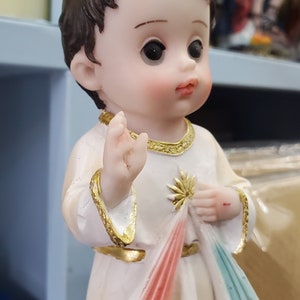 Puede incluir: Figura de un ni&ntilde;o peque&ntilde;o con cabello casta&ntilde;o, vistiendo una t&uacute;nica blanca con adornos dorados. El ni&ntilde;o sostiene una tela con detalles rojos y azules. La figura tiene una expresi&oacute;n serena y es un art&iacute;culo religioso.