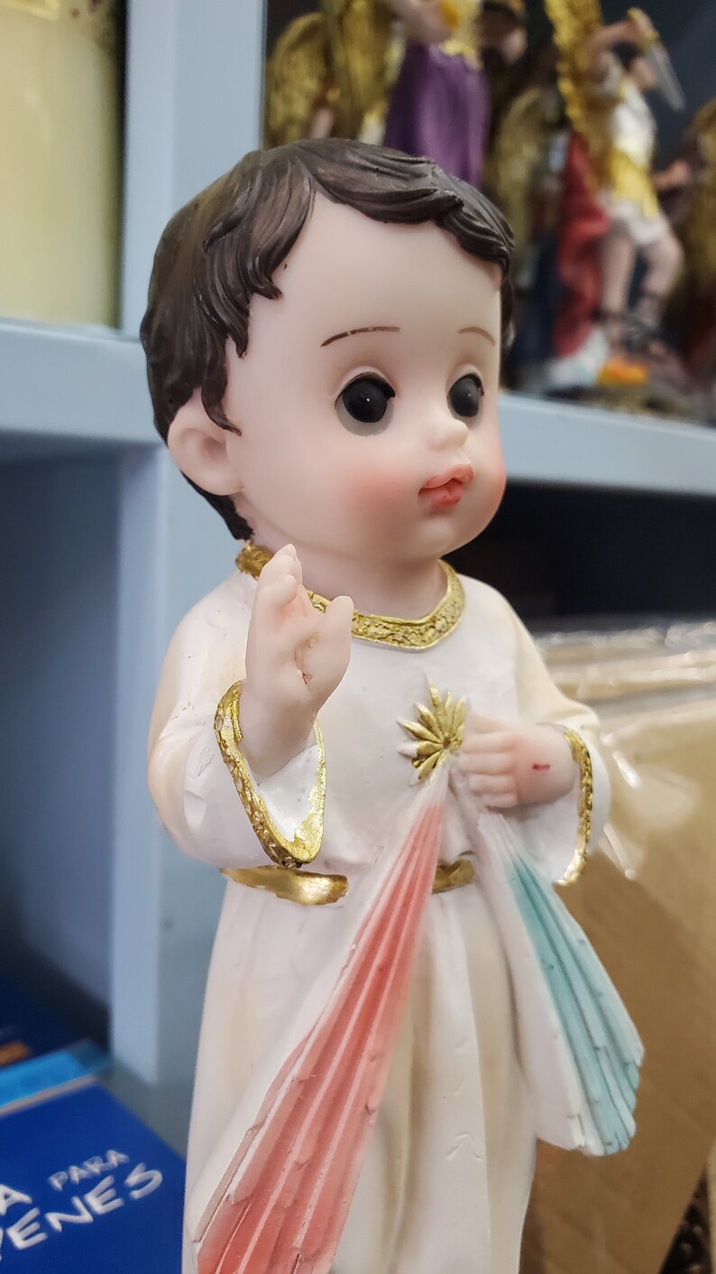 Puede incluir: Figura de un ni&ntilde;o peque&ntilde;o con cabello casta&ntilde;o, vistiendo una t&uacute;nica blanca con adornos dorados. El ni&ntilde;o sostiene una tela con detalles rojos y azules. La figura tiene una expresi&oacute;n serena y es un art&iacute;culo religioso.