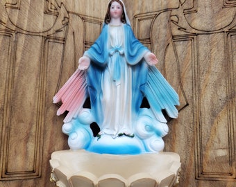 May include: A wooden wall shelf with a carved design of a cross and the word "INRI" on each side. A blue and white figurine of the Virgin Mary is placed on the shelf, standing on a cloud with a pink and blue halo. The figurine is holding her arms out in a gesture of welcome. Below the figurine is a beige shell-shaped dish with gold accents.