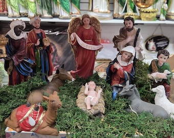 May include: A Christmas nativity scene with figurines of the Three Wise Men, an angel, Mary, Joseph, baby Jesus, and animals. The figures are brightly colored and set on a bed of green foliage.