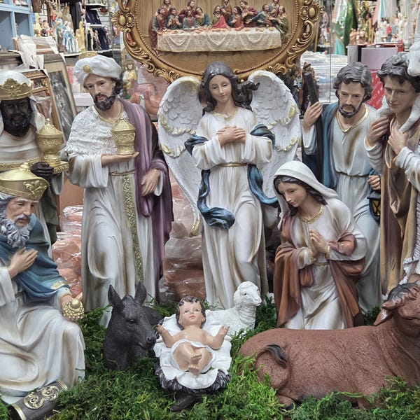 Może przedstawiać: Zestaw szopki bożonarodzeniowej z figurkami Świętej Rodziny, trzech mędrców, anioła, pasterza, osła, krowy i jagnięcia. Figurki są pomalowane w stonowanych odcieniach bieli, błękitu, brązu i złota, umieszczone na zielonej podstawie imitującej mech.