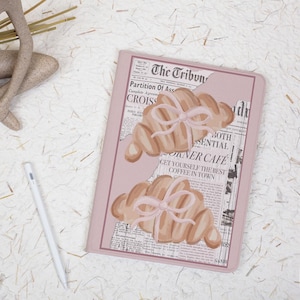 Op de afbeelding: Een roze notitieboek met illustraties van croissants vastgebonden met roze strikken. Het notitieboek heeft een achtergrond in krantenstijl met de tekst "The Tribune" en "Corner Cafe". Een witte styluspen ligt naast het notitieboek.