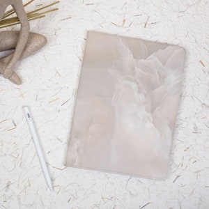 May include: A rectangular tablet case featuring a soft, beige and white marbled pattern with a smooth finish. Positioned next to the case is a slim, white digital stylus. The case has rounded corners and a clean, flat surface design.