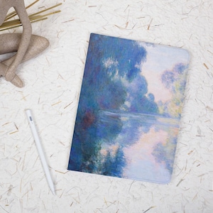 May include: A notebook featuring a blue and white impressionist painting of a waterscape. A white stylus and a decorative figurine are also visible. The notebook has a soft cover and is likely for writing or sketching.