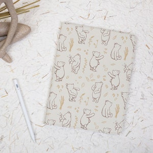 May include: A beige tablet case featuring a repeating pattern of brown bears and floral accents. A white stylus and a decorative statue are visible on a textured white surface.