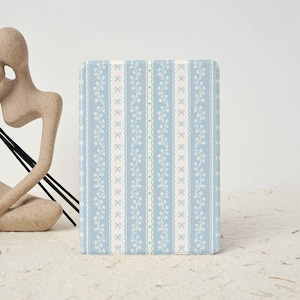 May include: A rectangular, light blue notebook with a repeating pattern of white bows and floral vines. The design is vertically striped with a cream-colored border. The notebook is sitting on a textured, off-white surface.