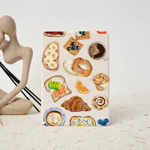 May include: A rectangular case featuring a watercolor illustration of breakfast foods. The design includes toast with avocado and salmon, waffles with blueberries, a bagel, a croissant, a muffin, and a cup of coffee.