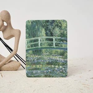 Puede incluir: Una impresión rectangular con una pintura impresionista de un puente arqueado verde sobre un estanque lleno de nenúfares, acompañada de una escultura abstracta beige de una figura humana con finos palos negros a un lado.