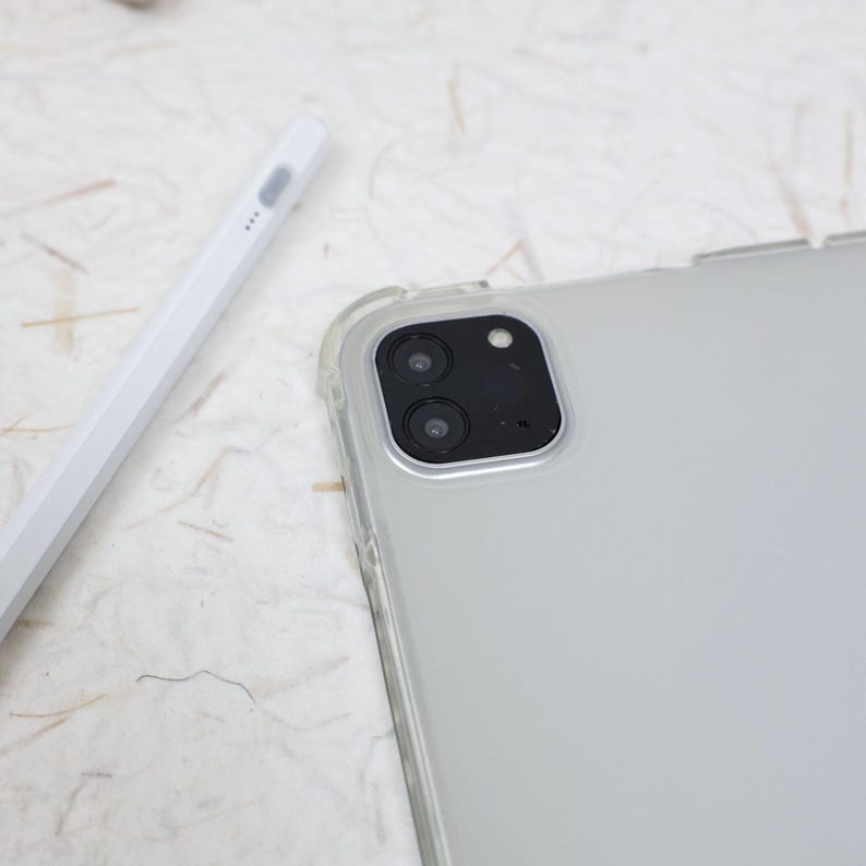 May include: A white digital stylus pen positioned next to a tablet device encased in a clear protective cover. The tablet features a prominent black square camera module housing two circular lenses and a small flash, set against a light-colored surface.