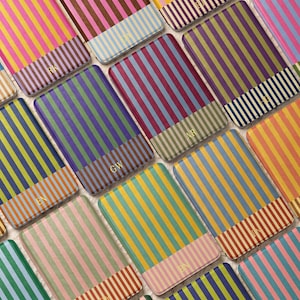May include: A collection of rectangular card holders featuring vertical stripes in various color combinations. Each holder has a distinct color palette and a two-letter monogram embossed in gold on the lower section of the front surface.