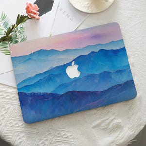 May include: A laptop case featuring a mountain landscape design in shades of blue, purple, and pink. The case has a white Apple logo in the center. The word "RENG" is visible on a white paper in the background.