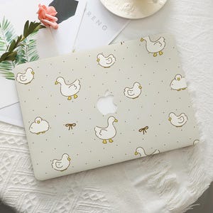 May include: A light gray laptop case featuring a pattern of white cartoon ducks with yellow beaks and feet, and small black dots. The case has a cutout for the Apple logo and small bow accents.