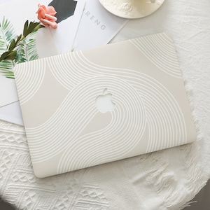 May include: A beige laptop featuring a white wave pattern. The Apple logo is visible. The laptop is on a white surface with a flower and greenery.