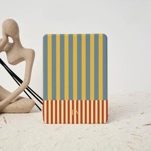 May include: A rectangular e-reader case featuring a vertical striped pattern. The top section has blue and yellow stripes, while the bottom section has red and cream stripes. The letters TN are centered in gold on the bottom section of the case.