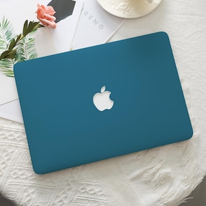May include: Teal laptop case featuring a white Apple logo. The rectangular case covers the top of the laptop. The background includes a white surface and decorative elements.