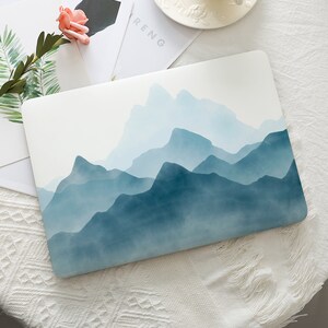 May include: A laptop case featuring a watercolor mountain range design in shades of blue. The design covers the top of the case, creating a serene and artistic aesthetic. The case is rectangular and appears to be made of a smooth material.