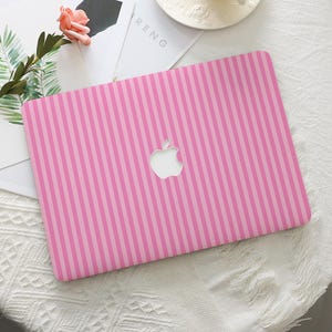 Può includere: Custodia rosa per laptop con un motivo a righe verticali in due tonalità di rosa. Il logo Apple è visibile al centro. La custodia è su una superficie bianca con un fiore e una rivista.