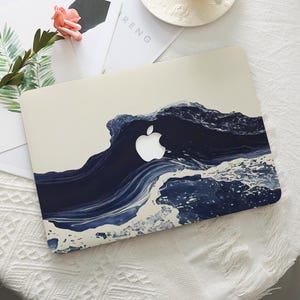 Puede incluir: Una funda para portátil con un diseño de olas en azul marino y blanco. El logotipo de Apple es visible en el centro. La funda tiene un acabado liso y está diseñada para proteger el portátil de arañazos y otros daños.