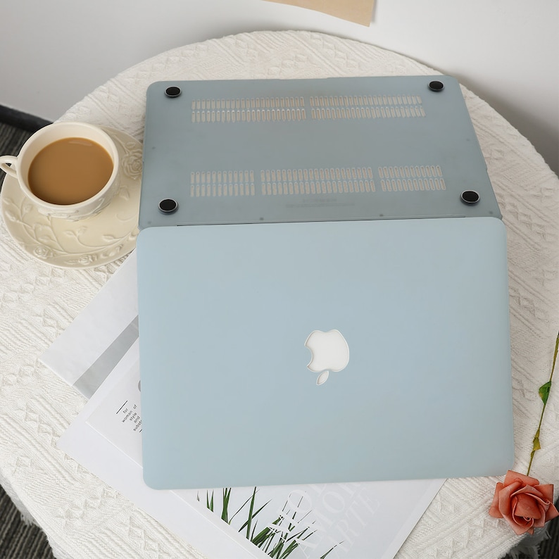 May include: A light blue laptop with the Apple logo, with a matching protective cover. The laptop is on a white table with a cup of coffee and some papers. The protective cover is a light gray color.