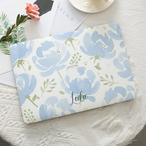 May include: A white laptop case featuring a watercolor floral design in shades of light blue and green. The case has a cutout for the Apple logo and the name "Leila" in a script font.