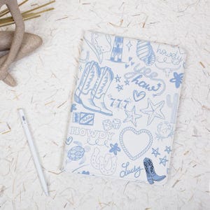May include: A white tablet case with a blue cowboy-themed design. The case features illustrations of cowboy boots, stars, hearts, and the word "Howdy". A white stylus pen is next to the case.