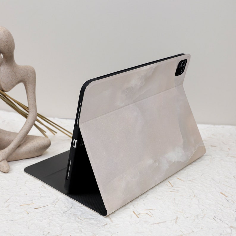 May include: A tablet case in a light beige marble pattern with a smooth finish. The case is folded into a stand position, revealing a black interior lining and a black frame that holds the tablet. The camera cutout is visible at the top right.