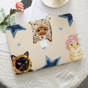 Könnte beinhalten: Eine beige Laptop-Hülle mit skurrilen Katzenillustrationen. Das Design umfasst eine Katze mit Blumenhaube, eine Katze mit Lockenwicklern und eine schwarze Katze mit gelber Kapuze. Denim-Sternakzente und ein weißes Apple-Logo sind ebenfalls vorhanden.