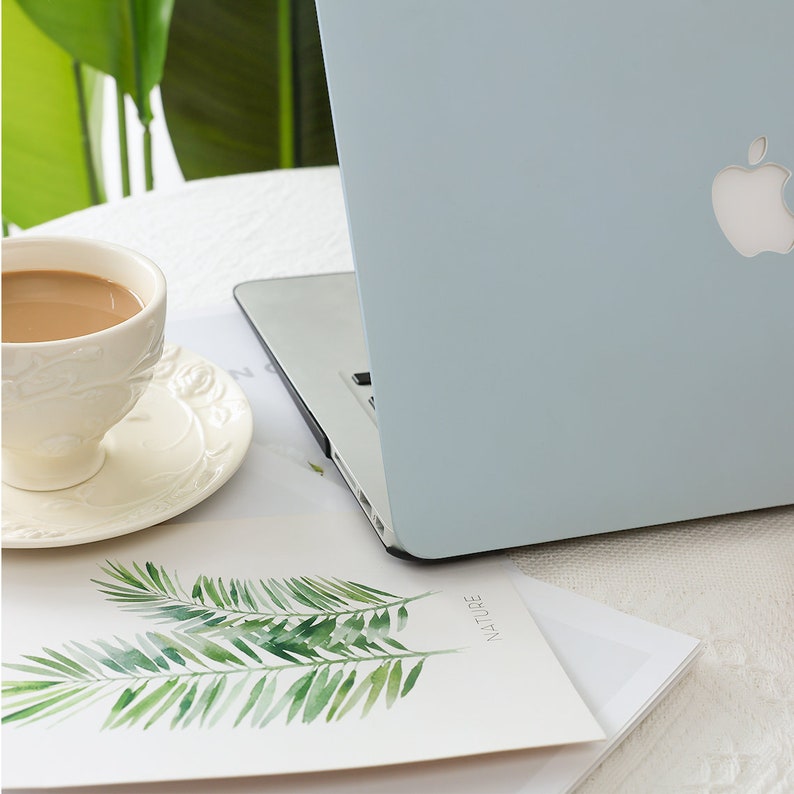 May include: A light blue laptop with a silver Apple logo sits on a white surface next to a cup of coffee and a saucer. A watercolor print of green palm leaves and the word "NATURE" is visible.